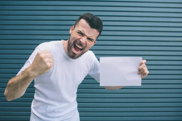 Brave man in white shirt is standing and posing on camera. He is holding fingers in fist. Also guy is holding piece of paper in other hand. He is screaming. Isolated on blue and striped background.