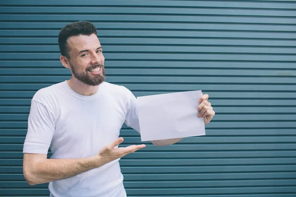 Nice and positive man iss standing and smiling. He is posing and looking at camera. Guy is holding piece of paper and poiting on it with hand. Isolated on striped and blue background. — Stock Photo, Image