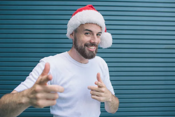 Positive guy with santa hat is looking on camera and smiling. He is pointing on camera. Bearded guy is happy. Isolated on striped and blue background. — Stock Photo, Image
