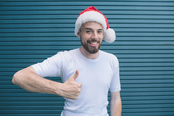 Good-looking and positive man is standing and looking on camera. He is showing his big thumb up. Also guy wears christmas hat. Isolated on striped and blue background. — Stock Photo, Image