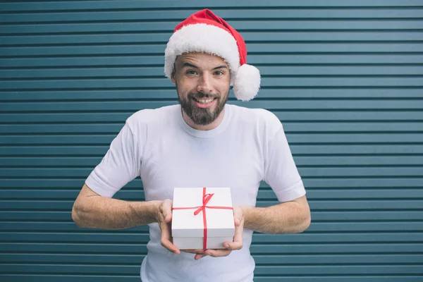 Delightful man in Christmas hat is standing and holding present in white cover. He is looking forward on camera. Guy is smiling. He is very positive. Isolated on striped and blue background. — Stock Photo, Image