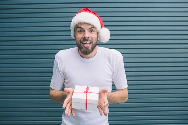 Excited bearded man is standing and holding present in hands. He is amazed. Man wears Christmas hat. Isolated on striped and blue background. — Stock Photo, Image