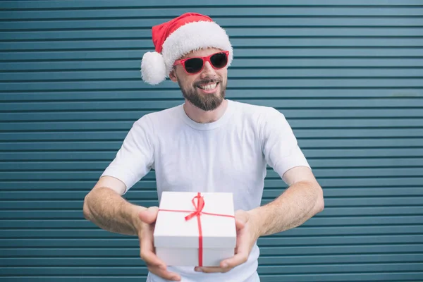 Bearded and happy man is standing and looking on camera. He wears Christmas hat and glasses. Man is smiling. Guy is holding present in hands. Isolated on striped and blue background. — Stock Photo, Image