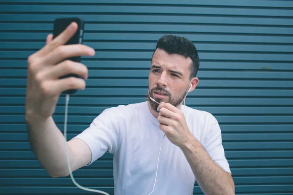 Guy is posing on camera. He is taking selfie and holding one hand on chin. Also man is listening to music. Isolated on striped and blue background. — Stock Photo, Image