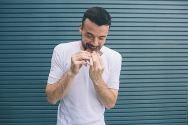 Esmer adam aşağı bakıyor ve parça çikolata ısırmak. Bu iki elinizle tutar. Çizgili ve mavi arka plan izole.