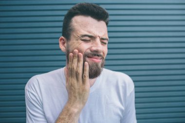 Genç adam ayakta ve elini yanağına tutuyor. Gözleri kapalı tutuyor. Adam acı hissetmiyor. Çizgili ve mavi arka plan izole.