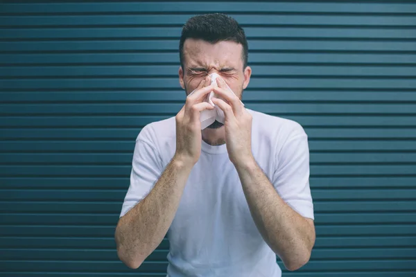 O tipo de camisa branca está a sofrer. Ele está cobrindo o nariz com guardanapo branco. O homem tosse e espirra. Isolado em fundo listrado e azul . — Fotografia de Stock