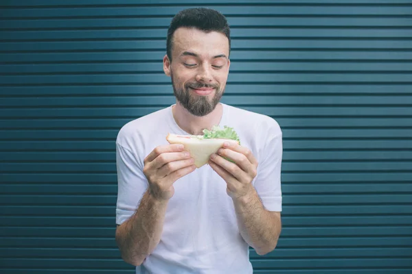 Glücklicher Kerl hält Sandwich in der Hand und betrachtet es. lächelt er. isoliert auf gestreiftem und blauem Hintergrund. — Stockfoto