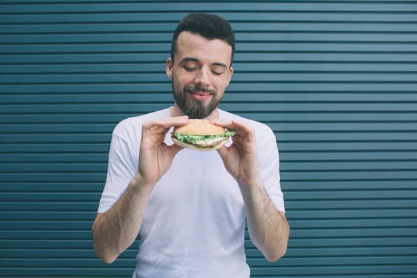 Der bärtige Mann hält Burger mit beiden Händen. Er sieht es an und lächelt. isoliert auf gestreiftem und blauem Hintergrund. — Stockfoto