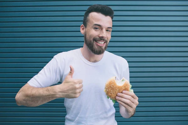 햄버거를 들고와 그의 큰 엄지손가락을 보여주는 남자의 그림. 그는 미소 하 고 카메라에. 줄무늬 및 파란 배경에 절연. — 스톡 사진