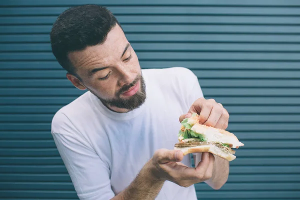 Brünetten und bärtigen Mann hält Sandwich und zeigt alle Gemüse, die zwischen zwei Brotstücken sind. Er schaut es sich an und lächelt ein wenig. isoliert auf gestreiftem und blauem Hintergrund. — Stockfoto