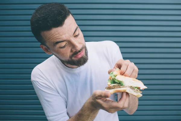 Morena e barbudo homem está segurando sanduíche e mostrando todos os vegetais que estão entre dois pedaços de pão. Ele está olhando para ele e sorrindo um pouco. Isolado em fundo listrado e azul . — Fotografia de Stock