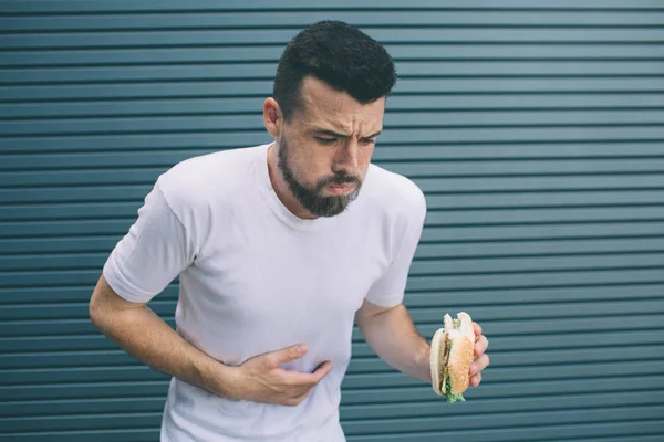 El tipo está enfermo. Él sostiene la hamburguesa en la mano y mira hacia abajo. El hombre va a vomitar. Aislado sobre fondo rayado y azul . — Foto de Stock
