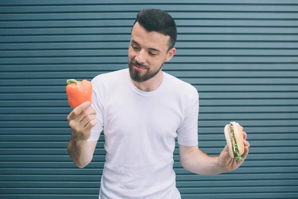 Nice guy is holding orange drink in one hand and burger in another one. He is looking at glass of drink. Isolated on striped and blue background. — Stock Photo, Image