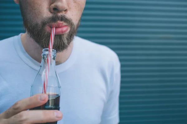 Cut view of man drinking coca-cola from small bottle through straw. He is holding it in one hand. Isolated on striped and blue background. — Stock Photo, Image