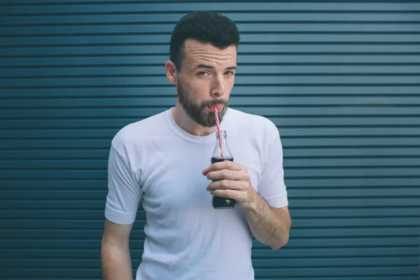 Vážný muž pije Coca-Colu z láhve brčkem. Dívá se na kameru. Izolované na pruhované a modré pozadí. — Stock fotografie