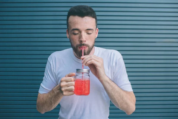 Rozkošný muž drží sklenici limonády a pití brčkem. On si užívá chvíle. Izolované na pruhované a modré pozadí. — Stock fotografie