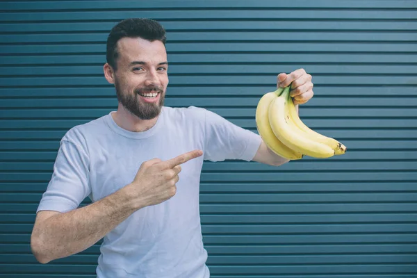 Happy guy is hlding bananas in one hand and pointing on them with another one. He is looking on camera and smiling. Isolated on striped and blue background.