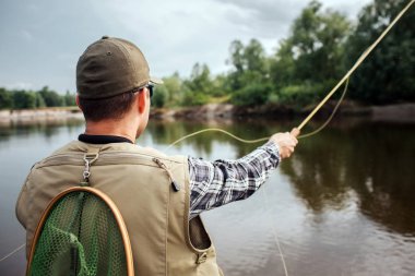 Adam ayakta ve düz üstünde su ve ağaçlar arıyorum. O yelek, kap ve gömlek giyer. Ayrıca adamın arkasından balık ağı var. Adam tek elle sinek çubuk tutar. Kaşık ileri atıyor