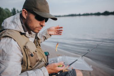 Güneş gözlüğü ile ciddi adam su ve aşağı bakıyor kenarında oturuyor. Yem kaşıkla dayadı. Sinek çubuk üzerinde kum yalan söylüyor.