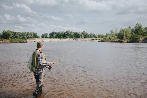 Una imagen del hombre de pie en el agua y la pesca. Está sosteniendo girando en las manos y mirando el agua. El tipo tiene una red de pesca en la espalda. Parece tranquilo y concentrado. . — Foto de Stock