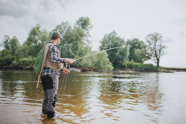 Adult stands in shallow and holding fly rod in hands. He is fishing. Guy is holding fly rod in one hand and part of spoon in the other one. Also man has a fishing net on the back.