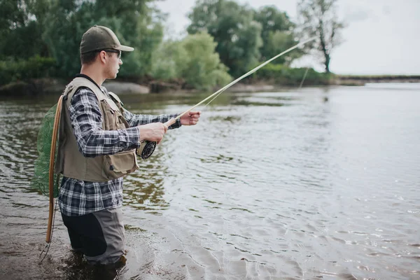 L'homme sérieux est debout dans l'eau et la pêche. Il a filé dans les mains et filet de pêche sur le dos. Le gars porte gilet, échassiers, chemise et casquette . — Photo