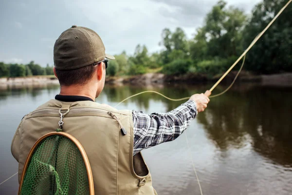 Adam ayakta ve düz üstünde su ve ağaçlar arıyorum. O yelek, kap ve gömlek giyer. Ayrıca adamın arkasından balık ağı var. Adam tek elle sinek çubuk tutar. Kaşık ileri atıyor