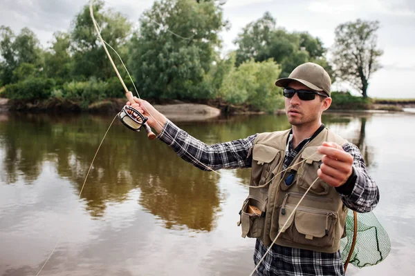 El hombre serio y concentrado se para en el agua y sostiene la pesca con mosca con carrete debajo de ella en una mano y parte de la cuchara en la otra. También el hombre tiene red de pesca en la espalda. Está pescando. . — Foto de Stock