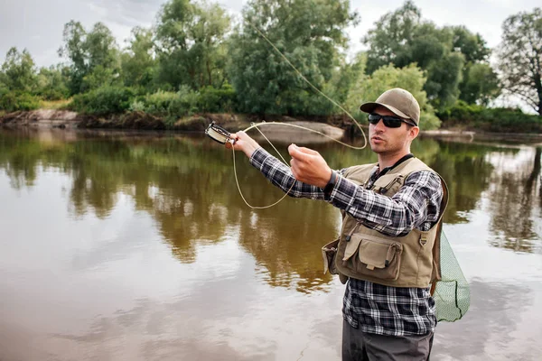 Un cuadro del hombre está parado en el agua y agitando con la barra de la mosca. Va a pescar. El tipo tiene una red de pesca en la espalda. Se ve serio y tranquilo. . — Foto de Stock