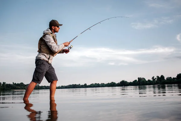 Активна людина стоїть на мілководді і рибалить. Він тримає мухоловку в руках. Чоловік крутиться навколо котушки, щоб зробити ложку коротшою і зловити рибу . — стокове фото