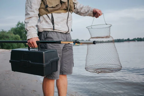 Uma foto de homem segurando rede de pesca e caixa preta de plástico nas mãos. Além disso, ele tem mosca rold no caminho certo. O tipo está na margem de Watr. . — Fotografia de Stock