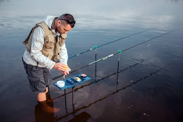 Un joven pescador está parado en el agua descalzo. Se inclina hacia una caja de plástico abierta con cebos artificiales. Hay dos varillas de mosca acostadas en ganchos . — Foto de Stock