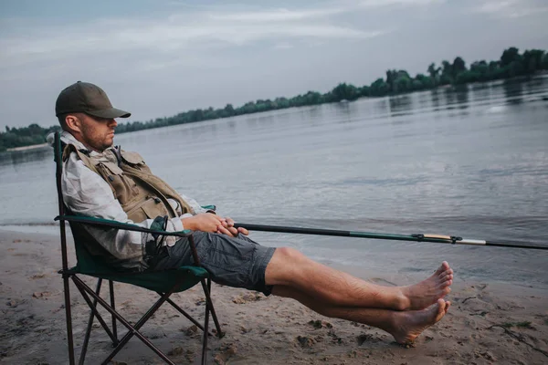 Fisherman está sentado en una silla plegable y durmiendo. Su gorra está sobre los ojos. El hombre sostiene la barra de mosca en las manos. Está pescando. Es de noche afuera. . — Foto de Stock