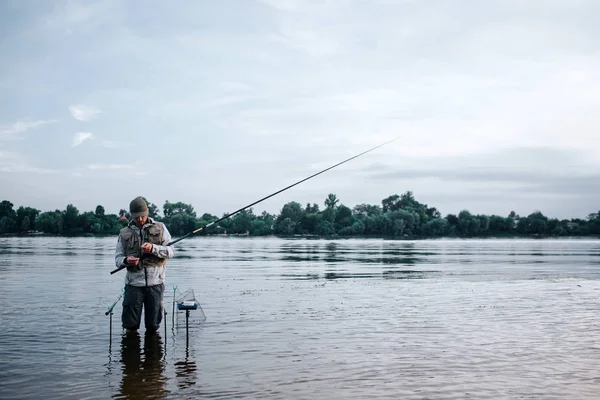 Genç balıkçı su ve sinek çubuk holding ayakta barefeet olduğunu. Öyle görünüyor. Adam kaşık ve yem ile çalışıyor. Yakınındaki bir ucu ile suya koymak kanca duruyor.