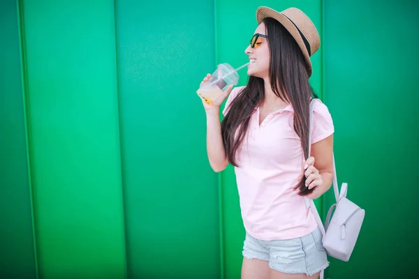 漂亮和愉快的女孩站在相机上摆姿势。她很期待。女孩正在用吸管喝水。女人拿着另一根头发。在绿色和条纹背景上隔离. — 图库照片