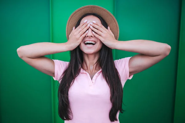 Glückliches Mädchen steht und hält die Augen mit den Händen geschlossen. , lächelt sie. Brünette trägt rosa Hemd und Strohhut. isoliert auf grün und gestreiftem Hintergrund. — Stockfoto