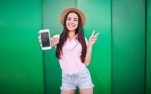 Nettes Mädchen steht und posiert vor der Kamera. Sie hält ein weißes Telefon in der Hand und zeigt ein Stück Symbol mit dem anderen. Frau lächelt. isoliert auf grün und gestreiftem Hintergrund. — Stockfoto