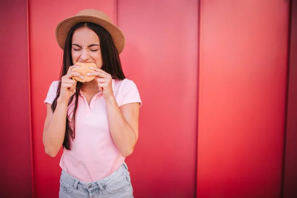 Una foto de una chica se para y muerde un trozo de rollo. Ella lo sostiene con ambas manos. La mujer mantiene los ojos cerrados. Está concentrada. Aislado sobre fondo rojo y rayado . — Foto de Stock