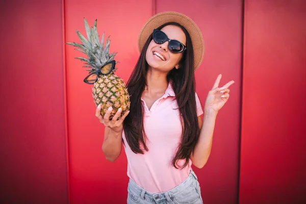 Vacker kvinna står och poserar på kameran. Hon visar symbolen bit med fingrarna. Kvinnan har ananas i höger hand. Både hon och frukten har glasögon. Isolatd på röd och randig bakgrund. — Stockfoto