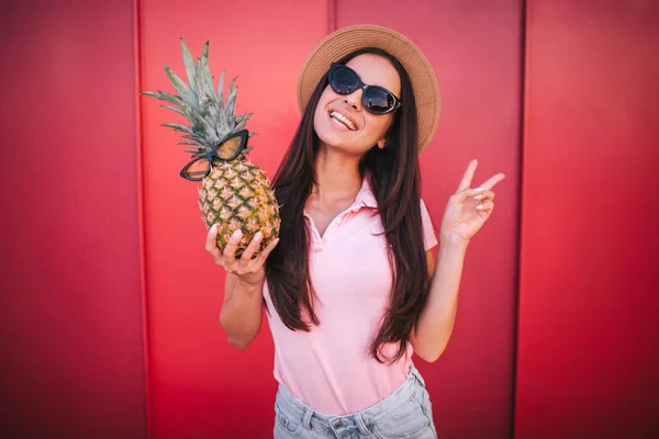 Schöne Frau steht und posiert vor der Kamera. zeigt sie das Stücksymbol mit ihren Fingern. Frau hat Ananas in der rechten Hand. Sowohl sie als auch die Früchte haben Gläser. isoliert auf rotem und gestreiftem Hintergrund. — Stockfoto