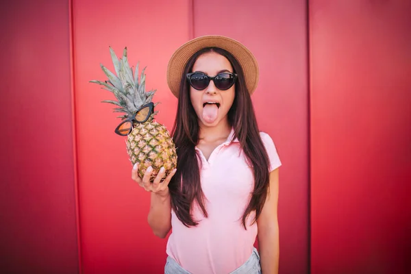 Lustiges Mädchen mit Sonnenbrille steht auf und zeigt ihre Zunge. Frau hält Ananas mit Gläsern. posiert sie vor der Kamera. isoliert auf gestreiftem und rotem Hintergrund. — Stockfoto