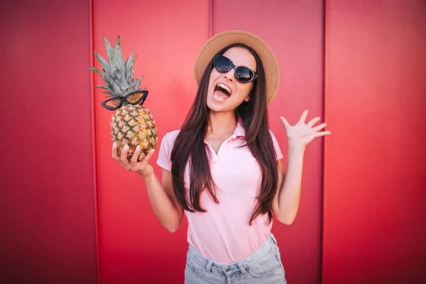 Glad och upphetsad brunett står och håller ananas i ena handen. Det har solglasögon. Flickan bär den också. Hon är förvånad. Isolerad på röd och randig bakgrund. — Stockfoto