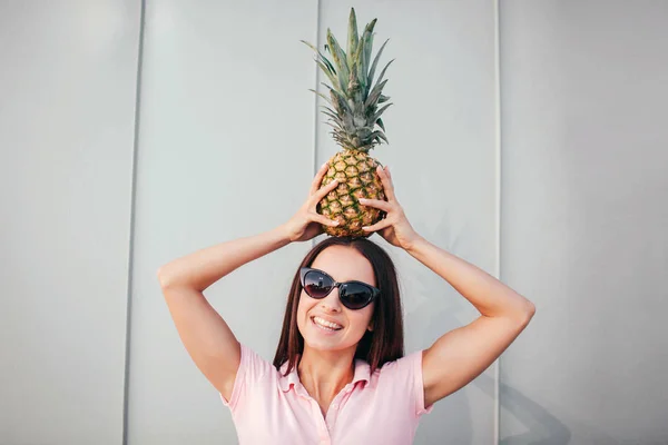 Positiv tjej i solglasögon står och håller ananas på huvudet. Hon håller den i balans med händerna. Flickan ser på camea och leenden. Isolerad på vit och randig bakgrund. — Stockfoto