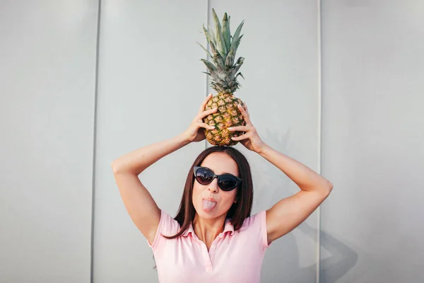 Lustiges erwachsenes Mädchen steht und hält Ananas auf ihrem Kopf. Sie trägt eine Sonnenbrille. Frau zeigt ihre Zunge. isoliert auf gestreiftem und weißem Hintergrund. — Stockfoto