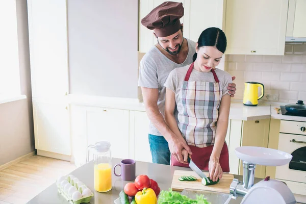 Small family is working together at kitchen. Man and woman cut cucumber together. Guy helps girl to do that. They prepare breakfast. — Stock Photo, Image
