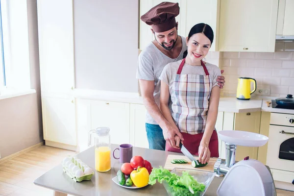 Beautiful couple stands at kitchen and wokr together. Girl looks at camera and smiles. She holds knife in one hand and cucumber in the other one. Guy helps her to cut vegetable. — Stock Photo, Image