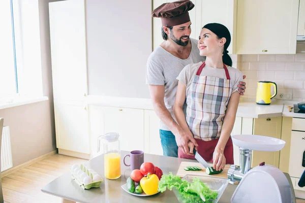 Delightful people are standing together at kitchen. They look at each other and smile. People look happy. Girl cuts cucumber with knife. Guy helps her to do that. — Stock Photo, Image