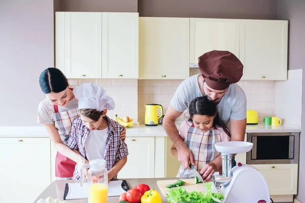 Προσεκτική τους γονείς στέκονται πίσω από τα παιδιά τους και βοηθώντας τους να κόψει τα λαχανικά. Που διδάσκουν τα παιδιά πώς να κόψει την ντομάτα και το αγγούρι σε ένα σωστό τρόπο. — Φωτογραφία Αρχείου