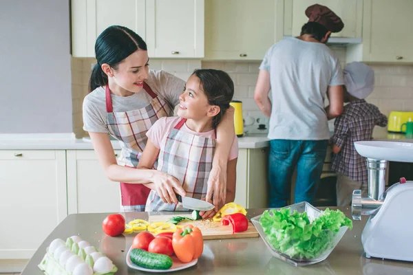 Позитивная девушка смотрит друг на друга и улыбается. Мать помогает дочери правильно резать овощи. Папа готовит еду с сыном на плите . — стоковое фото